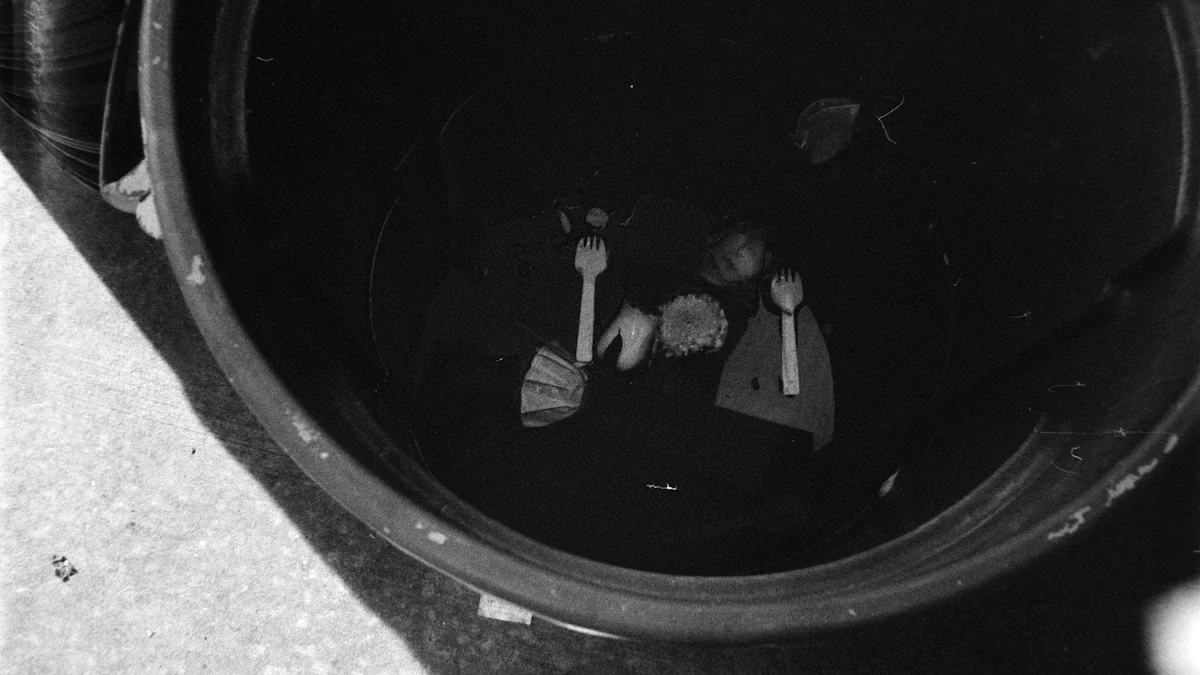 A dark trash bin containing discarded plastic cutlery and food waste items.