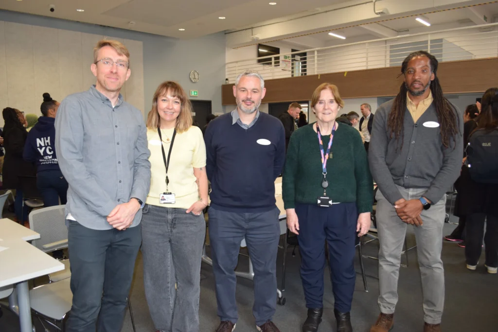 Five diverse professionals standing in a row and smiling for a group photo at an indoor event.