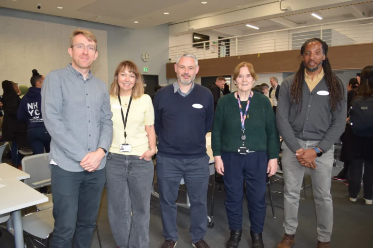 Five diverse professionals standing in a row and smiling for a group photo at an indoor event.