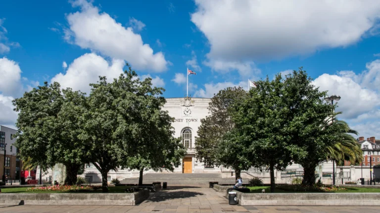 Art Deco style Hackney Town Hall behind lush green trees under a bright blue cloudy sky.