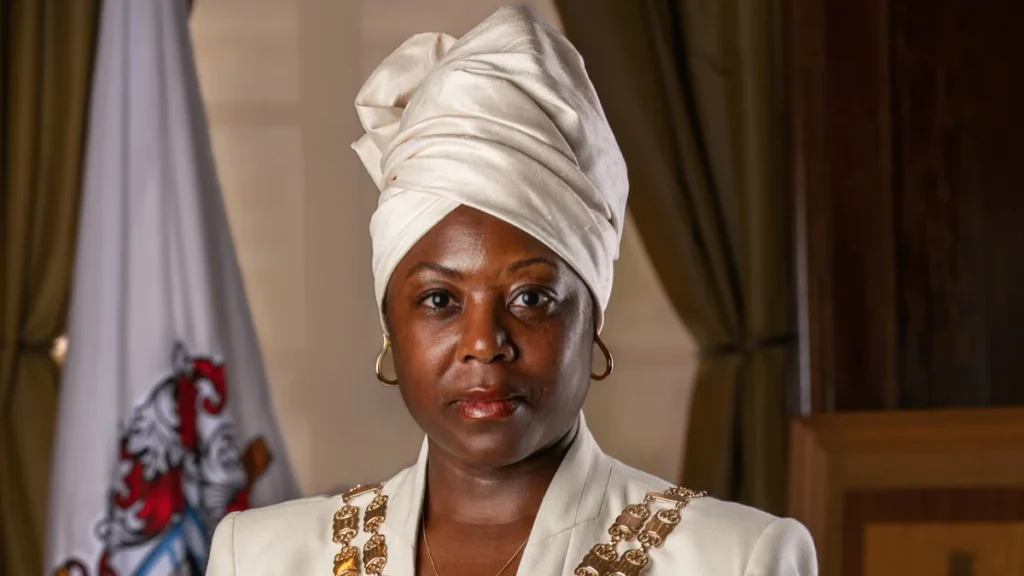 A Black woman wearing a white headwrap and formal chain of office looks forward.