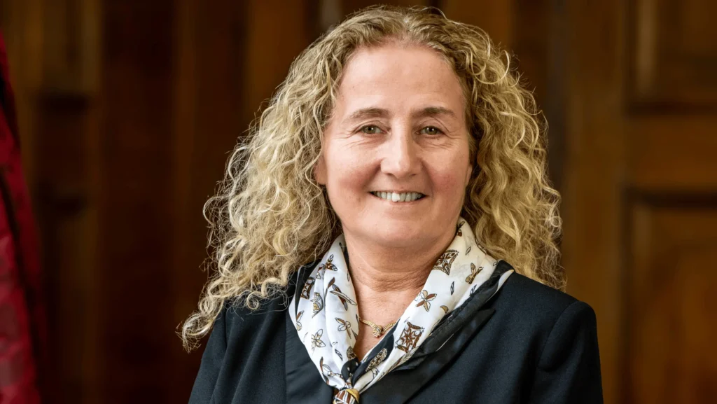 A smiling woman with curly blonde hair wearing a black blazer and a patterned scarf.