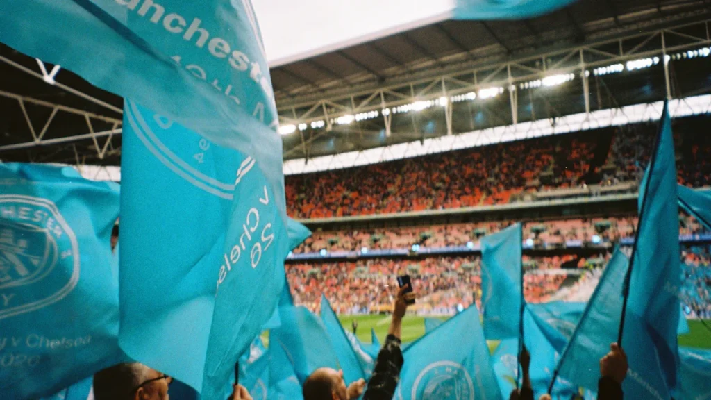 Manchester City fans waving light blue club flags inside a crowded football stadium.