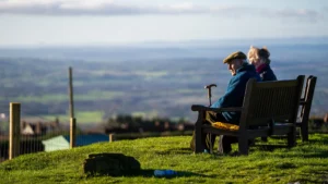 An elderly couple sits on a wooden bench overlooking a vast, scenic rolling countryside landscape.