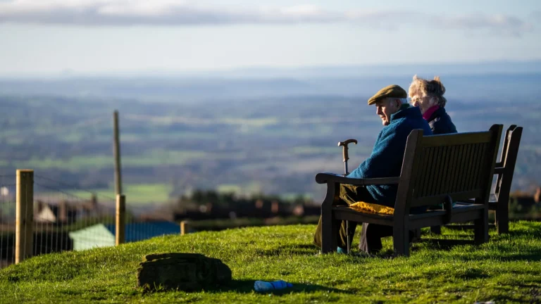 An elderly couple sits on a wooden bench overlooking a vast, scenic rolling countryside landscape.