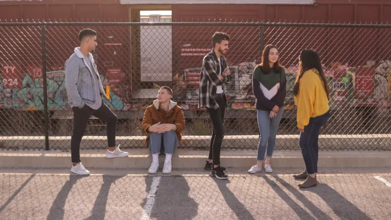 Five young people talking together while leaning against a chain link fence in the sun.