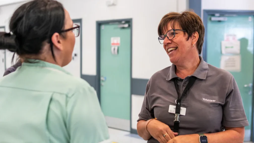 A friendly hospital volunteer in a grey polo shirt chatting with a nurse.