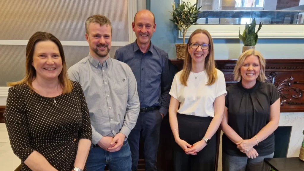 Five professional colleagues standing together and smiling in a bright office meeting room.
