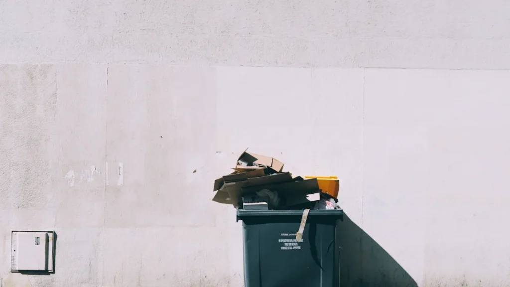 A black wheelie bin overflowing with cardboard boxes against a plain exterior wall.