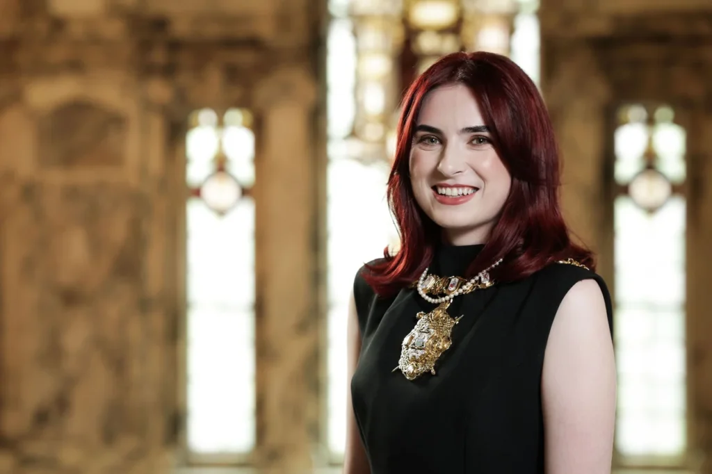 A smiling woman with red hair wearing a formal black dress and mayoral chain.