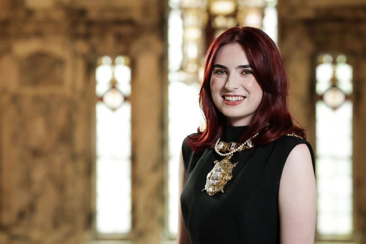 A smiling woman with red hair wearing a formal black dress and mayoral chain.