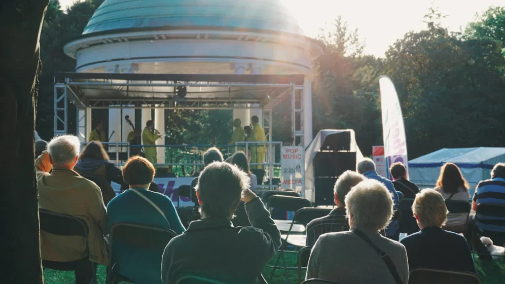 An audience watches a live band performing on a bandstand in a park.