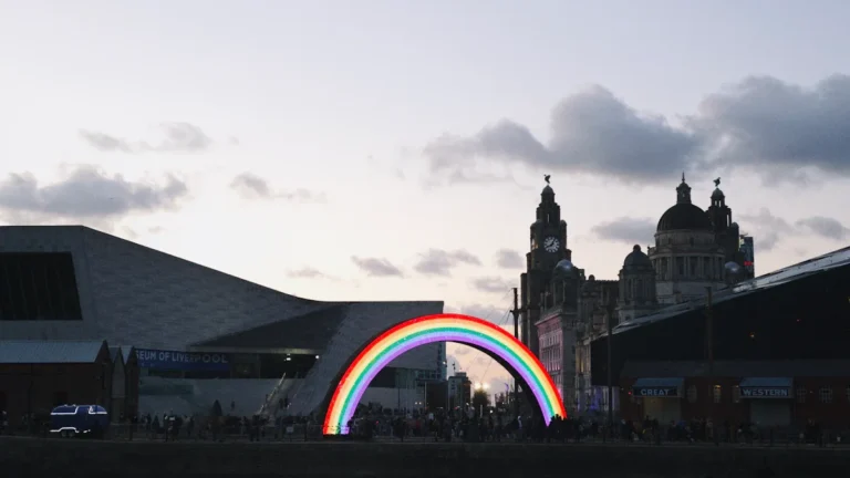 An illuminated rainbow arch installation set against the Liverpool city skyline at dusk.