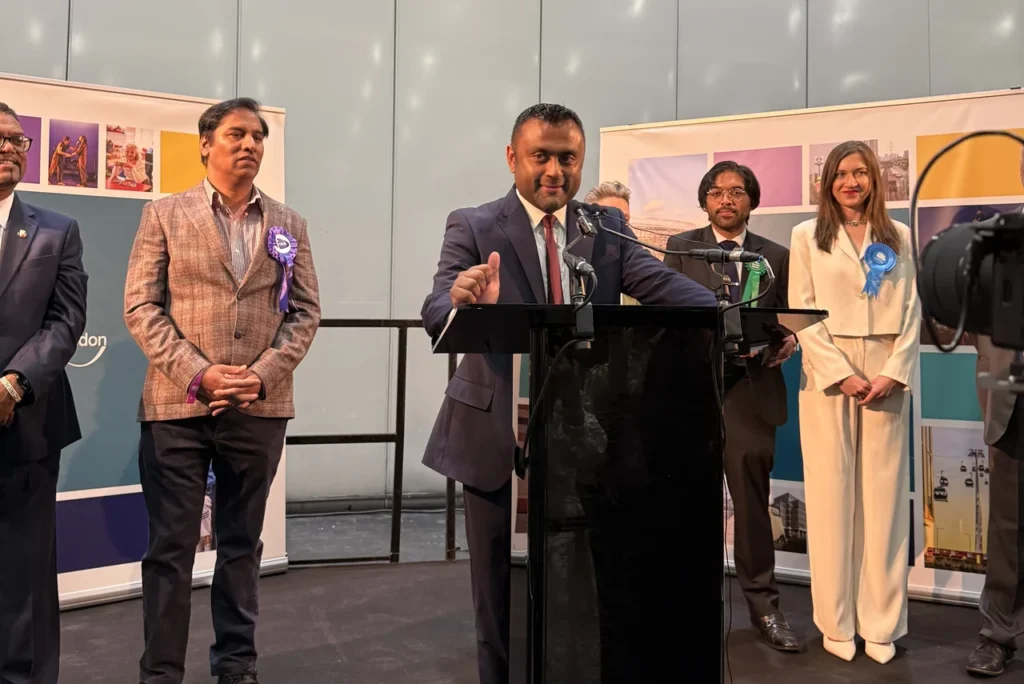 A man in a suit speaks at a podium with colleagues standing behind him.
