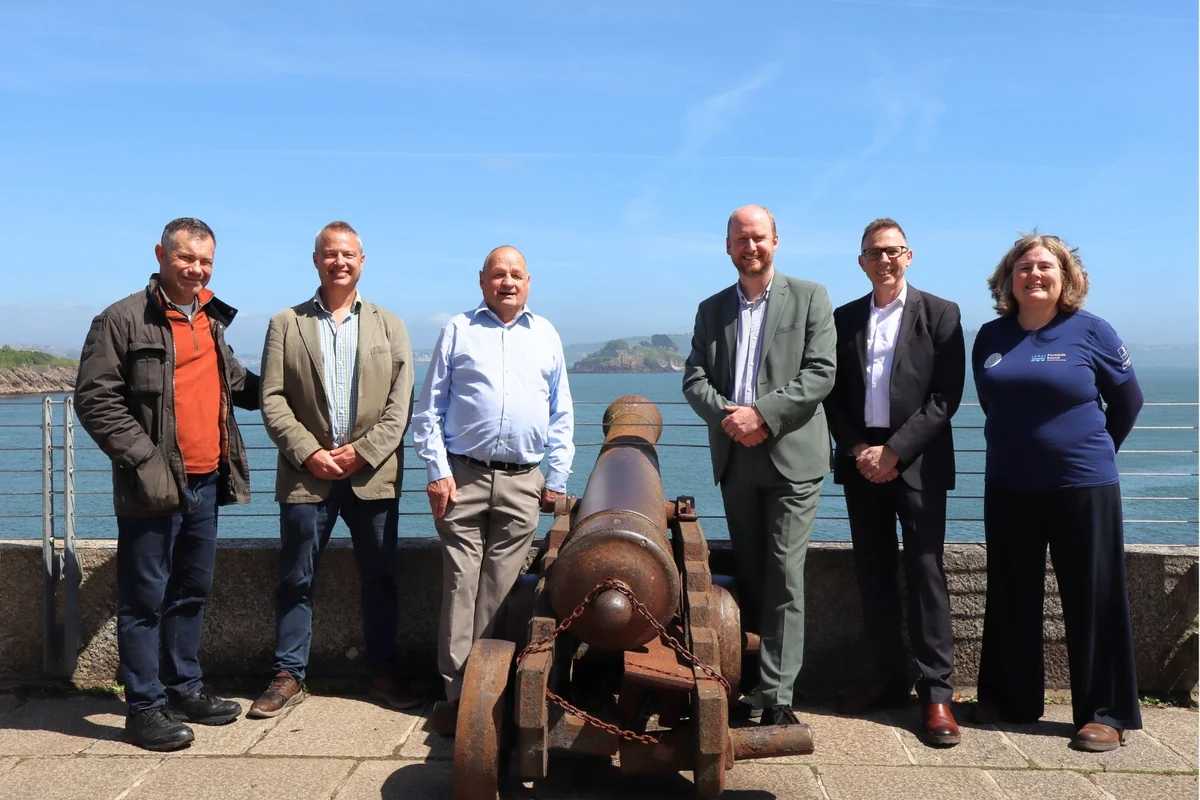 Group of six people posing outdoors with a historic rusty cannon overlooking the sea.