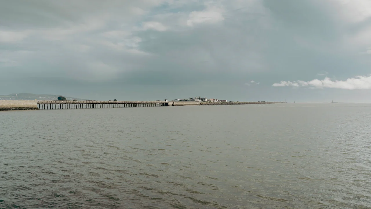 A calm, overcast coastal scene featuring a distant pier and shoreline buildings.