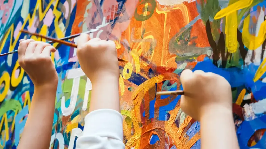 Close-up of children's hands painting a colorful mural with brushes on a wall.