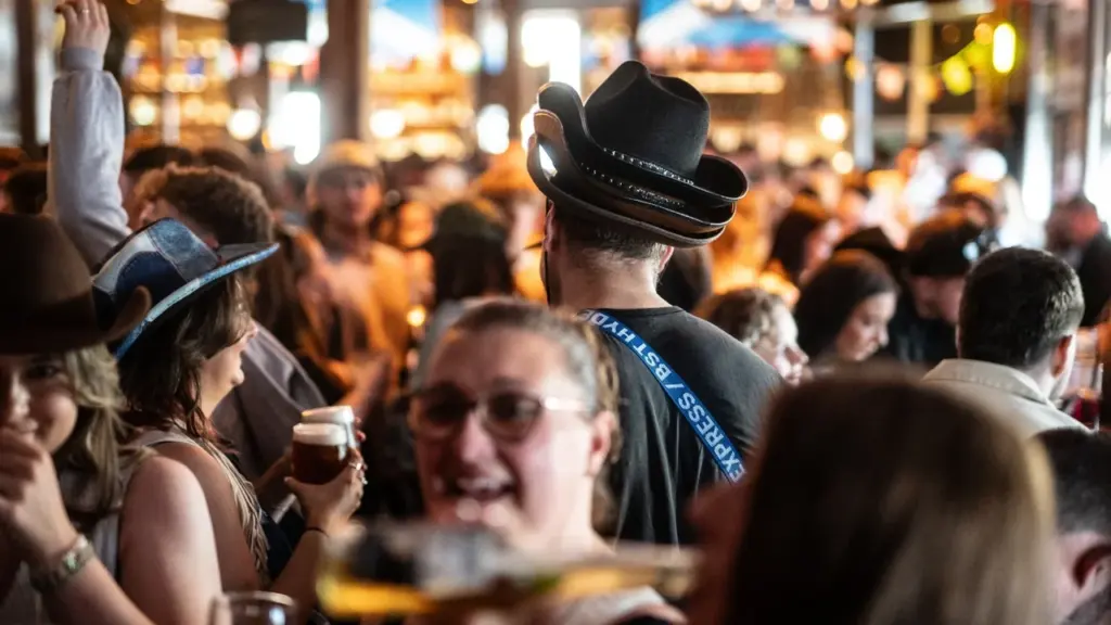 A crowded bar scene featuring people wearing hats, including a man in stacked cowboy hats.