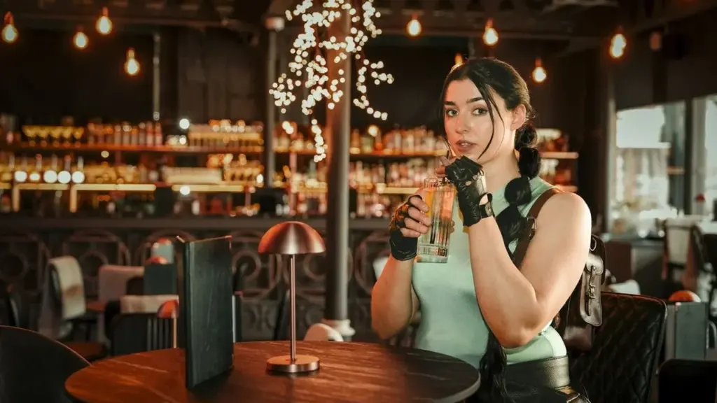 A woman dressed as Lara Croft drinks from a glass inside a dimly lit bar.