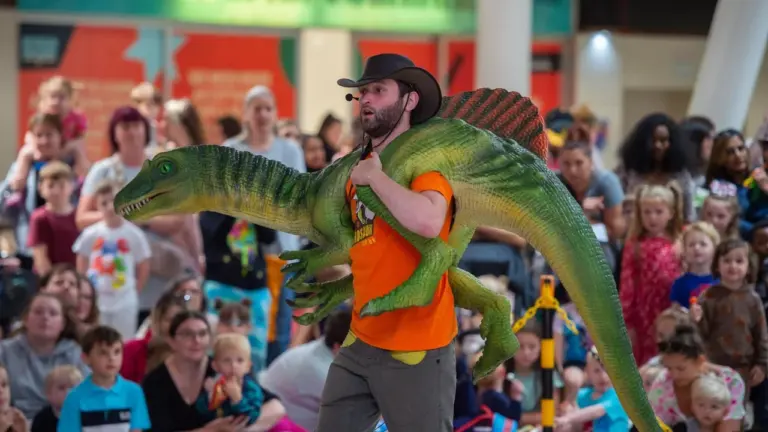 A man in a cowboy hat carries a green dinosaur puppet before a crowd.