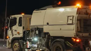 Street sweeper truck operating on city streets at night with lights on.