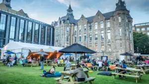 People sit at picnic tables and on beanbags in a grassy courtyard by historic buildings.