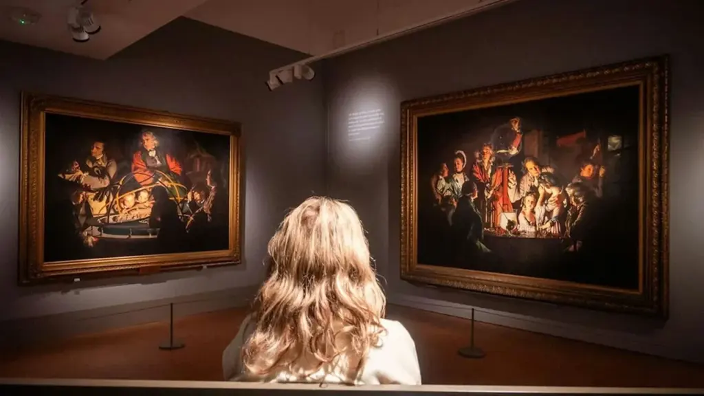 A woman with wavy blonde hair views two classic oil paintings in an art gallery.