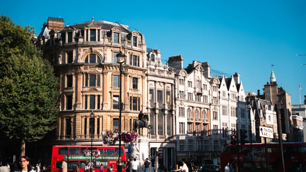 Classic red double-decker bus driving through a busy, sunny British city street scene.