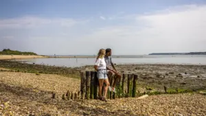 A young couple sits on weathered wooden posts along a sunny, pebble-covered coastal beach.