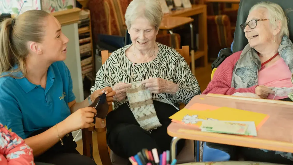 Caregiver assisting elderly residents with knitting in a warm, welcoming care home setting.