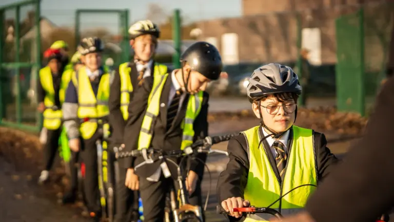 A line of school children in uniforms and high-visibility vests riding bicycles outdoors together.