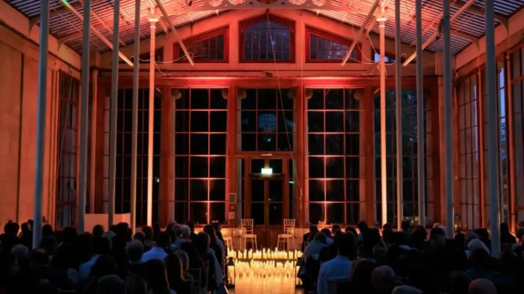 An audience watches a performance in a glass-walled building illuminated by warm red lighting.