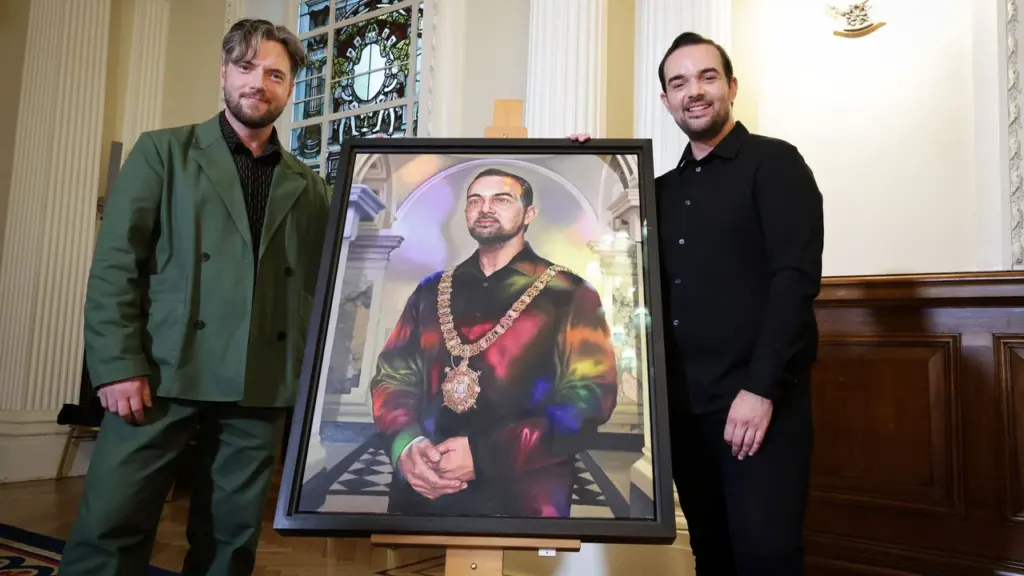 Two men stand beside an easel displaying a framed portrait of a mayoral figure.