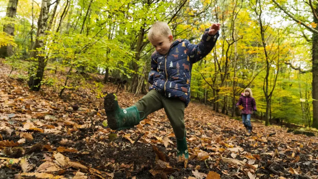 A young boy in a patterned jacket kicks mud in a forest with fallen leaves.