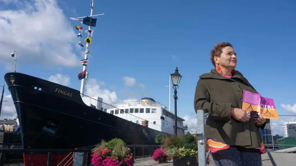 A woman stands holding a book in front of the Fingal ship moored outdoors.