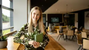 A smiling blonde woman wearing a green leaf-patterned blouse holding a green notebook indoors.