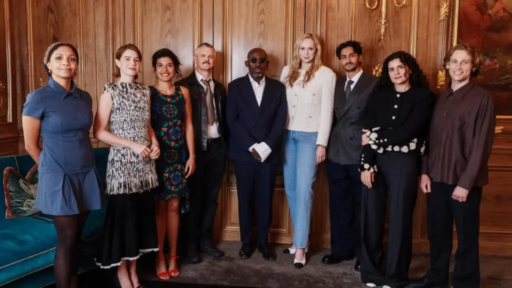 A diverse group of nine well-dressed actors standing together against a wooden paneled wall.